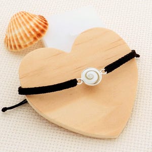 Op de afbeelding: Een zwart koordarmbandje met een cirkelvormige, wit en zilveren hanger met een spiraalvormig ontwerp. De armband wordt getoond op een houten hartvormig oppervlak, met een zeeschelp op de achtergrond.