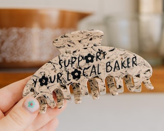 May include: A beige and black marbled hair claw clip featuring the black text "SUPPORT YOUR LOCAL BAKER" printed across the front. The clip has a series of interlocking teeth along the bottom edge and a spring-loaded hinge mechanism at the top.
