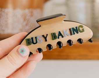 May include: A beige hair claw clip with a black top edge and black teeth. The text BUSY BAKING is printed on the side in dark green and black letters. The clip has a curved, semi-circular shape and a smooth, glossy finish.