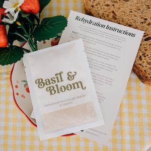 May include: A packet of dehydrated sourdough starter, "Basil & Bloom", containing 10 grams. The image shows rehydration instructions, a slice of bread, and a floral arrangement with strawberries and white flowers, on a yellow and white checked cloth.
