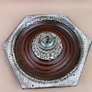 Op de afbeelding: Een zeshoekige keramische lampvoet met een bruin en wit gespikkelde glazuur. De voet heeft concentrische bruine ringen en een centrale metalen lamphouder. Het algehele ontwerp is retro en modern.