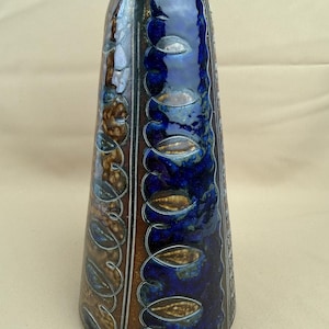 Op de afbeelding: Een hoge keramische vaas met een uitlopende bovenkant. De vaas heeft een glanzend, donkerblauw glazuur met bruine en witte accenten. Decoratieve blad- en wijnstokpatronen sieren de zijkanten. De vaas is ongeveer 25 cm hoog.