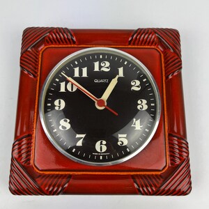 Könnte beinhalten: Eine quadratische, rote Quarzuhr mit schwarzem Zifferblatt und weißen Ziffern. Die Uhr hat einen silbernen Rand und rote Zeiger. Das Zifferblatt zeigt "QUARTZ" und "MADE IN GERMANY". Der Rahmen der Uhr hat ein strukturiertes Design.