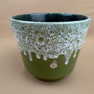 Può includere: Un vaso in ceramica con una glassa bianca e verde strutturata. Il bordo superiore è verde scuro, che sfuma in un motivo bianco maculato che gocciola verso una base verde unita. Il vaso ha un piccolo logo sul davanti.