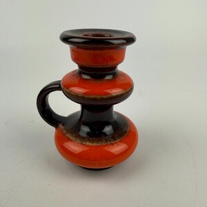 Könnte beinhalten: Ein brauner und orangefarbener Kerzenhalter aus Keramik mit einem Griff. Der Halter hat eine breite Basis und eine schmale Oberseite mit einem Ring zum Halten einer Kerze.