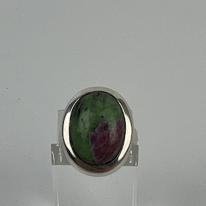Könnte beinhalten: Ein Silberring mit einem ovalen Zoisitstein. Der Stein zeigt eine Mischung aus grünen und kastanienbraunen Farben. Der Ring wird auf einem transparenten Ständer präsentiert, der den Schmuck hervorhebt.