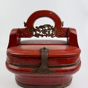 Può includere: Una scatola di legno rossa con una maniglia intagliata e dettagli in ottone. La scatola ha un coperchio incernierato e una chiusura in metallo. La scatola è in buone condizioni e ha un aspetto vintage.