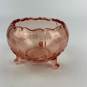 Könnte beinhalten: Eine rosa Glasschale mit gewelltem Rand und drei kleinen Füßen. Die Schale hat ein Blumendesign, das in das Glas geätzt ist. Die Schale ist hellrosa und wahrscheinlich vintage.