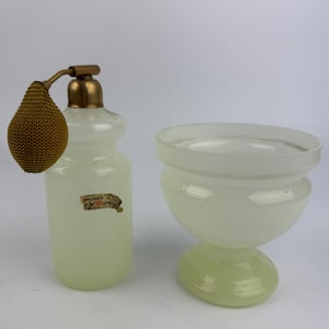 Può includere: Un flacone di profumo vintage con un vaporizzatore color oro e una ciotola di vetro abbinata. La bottiglia è di colore verde pallido con un'etichetta che dice "ristable de Murano". La ciotola è anch'essa verde pallido e ha una forma arrotondata.