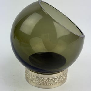 Può includere: Una moderna ciotola in vetro verde oliva con bordo argentato, posta su una base color argento con un motivo floreale ornamentale ripetuto. La ciotola ha un'apertura angolata.