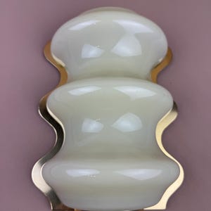 Könnte beinhalten: Eine Wandleuchte mit einer geschichteten, organischen Form. Die Leuchte hat einen cremeweißen Glasschirm und eine goldfarbene Metallrückseite. Das Design ist symmetrisch, mit einem bauchigen oberen und unteren Teil, die durch eine schmalere Mitte verbunden sind.