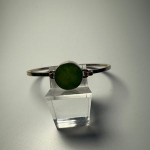 Könnte beinhalten: Ein silbernes Armreif-Armband mit einem runden grünen Stein in der Mitte.