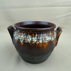 Könnte beinhalten: Eine braune Keramikschale mit zwei Griffen. Die Schale hat einen strukturierten, dunkelbraunen Boden und einen helleren braunen Mittelteil mit weißen und gelben Akzenten. Der Innenraum ist dunkelbraun.