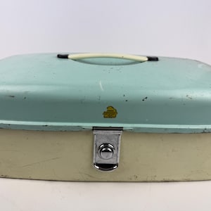 Könnte beinhalten: Eine vintage Brotdose aus Metall in Hellblau und Creme mit einem schwarzen Griff und einem silbernen Verschluss. Die Dose hat ein Etikett mit der Aufschrift "Bake-A-Way" in gelben Buchstaben.