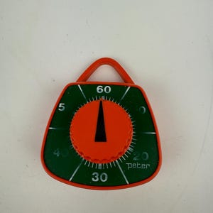 Könnte beinhalten: Ein grüner und orangefarbener Küchentimer mit einem schwarzen Zeiger und dem Wort "peter" auf dem Zifferblatt. Der Timer hat Zahlen von 5 bis 60.