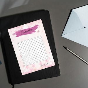 May include: A word search puzzle with the word "Surprise" in pink, on a black notebook. A light blue envelope and a pencil are on the grey surface. The puzzle has the text "En 5 mots..."