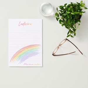 Op de afbeelding: Een witte notitieblok met gelinieerde pagina's, met een aquarel regenboogontwerp in pastelkleuren. Het notitieblok heeft de tekst "Ludivine" en "Ma vie en couleur" in cursief schrift. Een bril en een kleine potplant zijn ook zichtbaar.