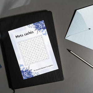May include: A black notebook with a French word search puzzle titled "Mots cachés", decorated with blue floral designs. A light blue envelope and a pencil are also present, suggesting a stationery or gift theme.