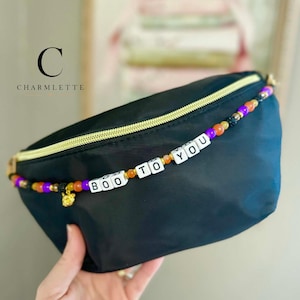 May include: A black fanny pack featuring a gold zipper and a decorative beaded strap across the front. The strap includes orange, purple, and black beads with white square letter beads spelling out the phrase BOO TO YOU and a small gold charm.