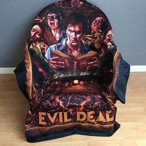 Pu&ograve; includere: Coperta in pile nera e arancione con una grafica di un uomo che legge un libro circondato da zombie. Il testo "Evil Dead" &egrave; stampato sul fondo della coperta.