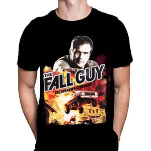 May include: Black t-shirt featuring a movie poster design. The graphic includes a man's portrait, the words "THE FALL GUY" in white, and a scene of a truck jumping over an explosion. The text "THE UNKNOWN STUNTMAN" is also present.