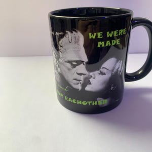 May include: Black ceramic mug featuring a grayscale image of Frankenstein's monster and his bride. The mug has the text "WE WERE MADE FOR EACH OTHER" in green lettering. The mug has a black handle.