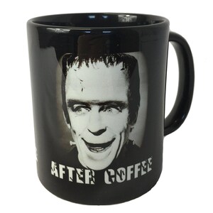Op de afbeelding: Zwarte koffiemok met een zwart-wit afbeelding van een lachend monster. De mok heeft de woorden "AFTER COFFEE" in witte blokletters. De mok heeft een zwart handvat.