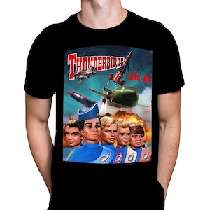 Peut inclure: T-shirt noir avec un graphisme coloré de la série Thunderbirds. Le motif comprend le texte "Thunderbirds Are Go" avec des images de fusées, d'avions et des personnages de la série. Le t-shirt est à col rond classique.