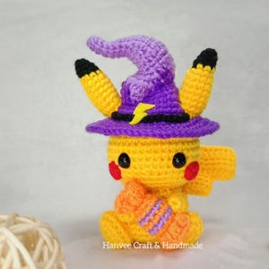 May include: A handmade crochet Pikachu figure wearing a purple witch hat with a yellow lightning bolt. The figure is yellow with black-tipped ears, red cheeks, and holding an orange and purple striped item. Text on the bottom reads "Hanvee Craft & Handmade."