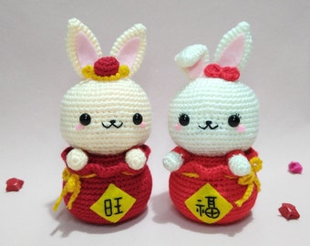 May include: Two white crocheted bunny plushes wearing red crocheted outfits with Chinese characters. The bunny on the left has a red and yellow crocheted hat. The bunny on the right has a red crocheted bow.