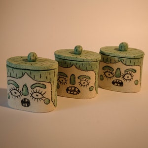 Op de afbeelding: Drie keramische containers met groene deksels en beschilderde gezichten. Elke container heeft een andere gezichtsuitdrukking.