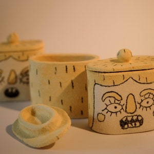 Op de afbeelding: Drie keramische containers met deksels, elk met een ander handgetekend gezicht. De containers zijn beige met zwarte contouren en details. Eén container heeft een eenvoudig gezicht met een neus en ogen, een andere heeft een gezicht met een neus en ogen, en de derde container heeft een gezicht met een neus, ogen en een mond.