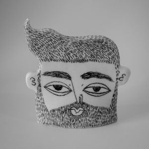 May include: A white ceramic sculpture of a person's face with a black and white drawn design. The sculpture has a beard, eyebrows, and eyes. The hair is drawn in a spiky style.