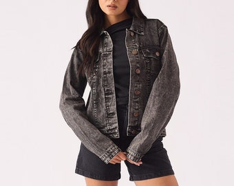 May include: A black denim jacket with a button closure and a faded look. The jacket has a relaxed fit and is worn over a black long-sleeved shirt. The model is wearing black shorts and black heeled sandals.