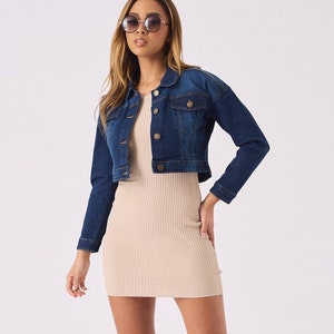 Op de afbeelding: Een vrouw draagt een donkerblauw denim jack met een lichtbeige geribbelde gebreide mini-jurk en bruine sandalen.