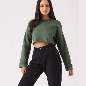 May include: A woman is wearing a green cropped sweatshirt and black high-waisted jeans. She is wearing black sneakers.