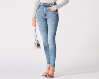 Könnte beinhalten: Eine Frau trägt ein hellgraues Langarmshirt mit asymmetrischem Ausschnitt und hellblaue Skinny Jeans. Sie trägt eine kleine silberne Handtasche und eine silberne Sonnenbrille.