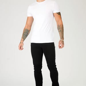 K&ouml;nnte beinhalten: Ein Mann tr&auml;gt ein wei&szlig;es T-Shirt und eine schwarze Jeans. Er steht mit seinen Armen an seinen Seiten und schaut nach links.