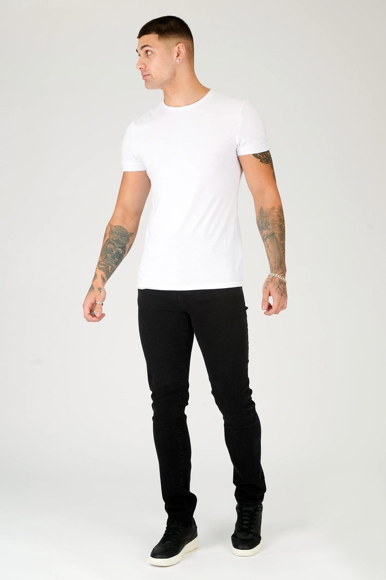 K&ouml;nnte beinhalten: Ein Mann tr&auml;gt ein wei&szlig;es T-Shirt und eine schwarze Jeans. Er steht mit seinen Armen an seinen Seiten und schaut nach links.