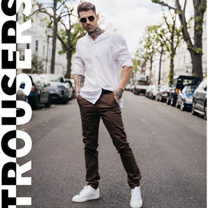 May include: A person wearing a white shirt and brown trousers, with white trainers and sunglasses. The word "TROUSERS" is displayed in large white letters on the left side of the image. The person is standing on a street with parked cars.