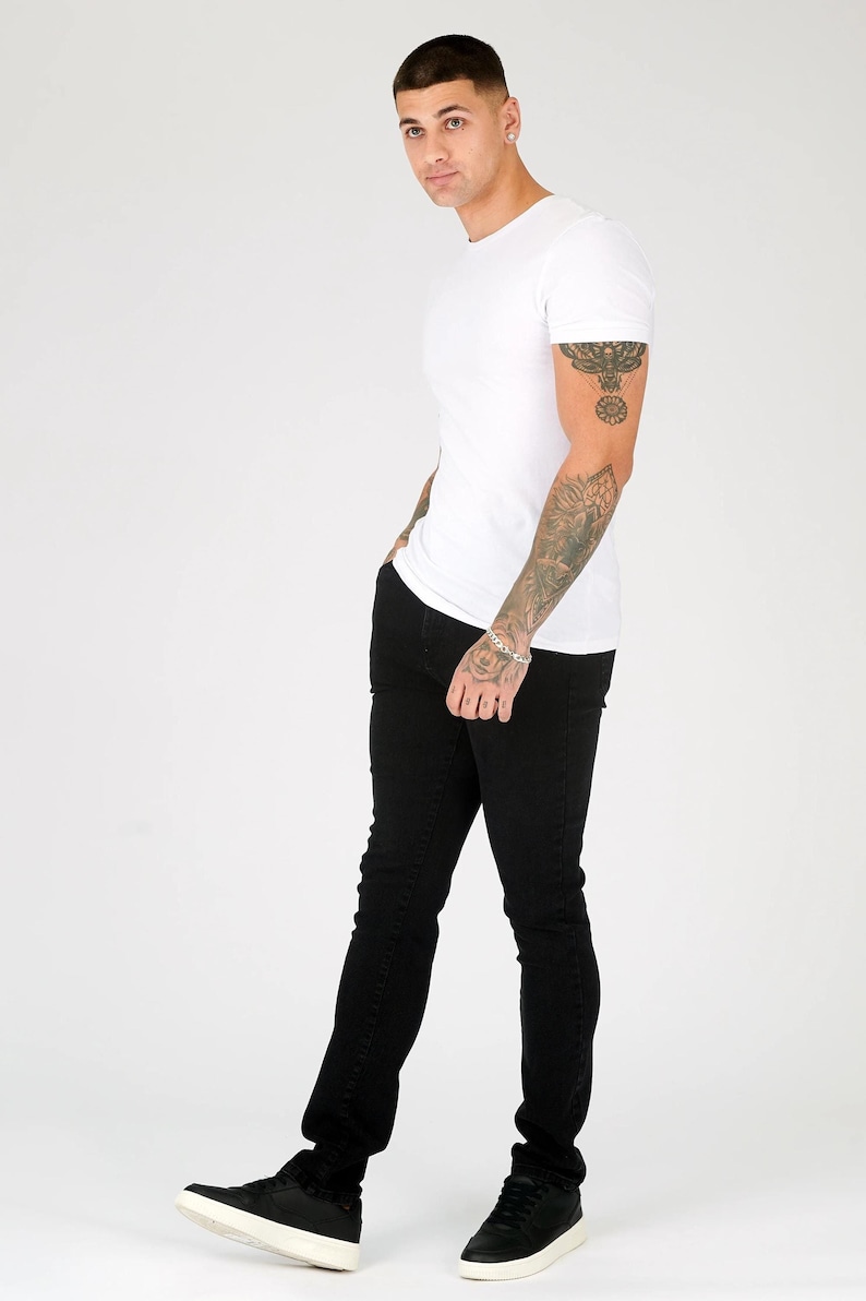 K&ouml;nnte beinhalten: Ein Mann tr&auml;gt ein wei&szlig;es T-Shirt und eine schwarze Jeans. Er steht mit der rechten Hand in der Tasche und der linken Hand auf der H&uuml;fte. Er tr&auml;gt schwarze Turnschuhe.