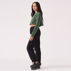 May include: A woman wearing a green cropped sweatshirt and black high-waisted jeans. She is wearing black sneakers.