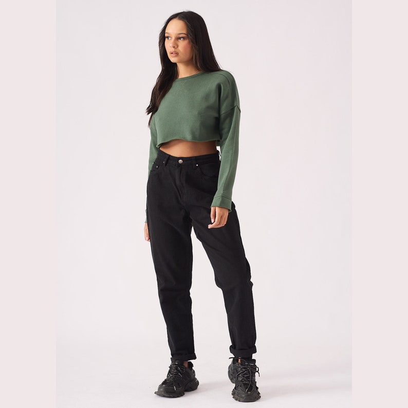 May include: A woman is wearing a green cropped sweatshirt and black high-waisted jeans. She is wearing black sneakers.