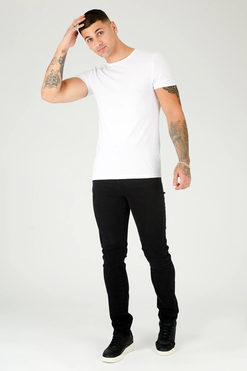 K&ouml;nnte beinhalten: Ein Mann tr&auml;gt ein wei&szlig;es T-Shirt und eine schwarze Jeans. Er steht mit der Hand am Kopf und schaut in die Kamera.