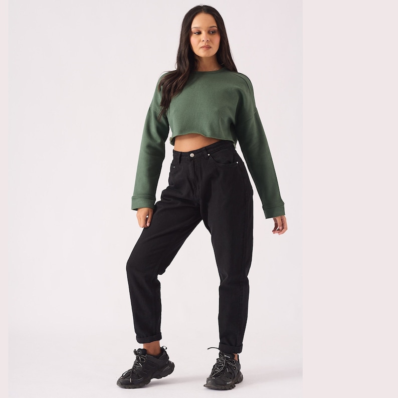 May include: A woman is wearing a green cropped sweatshirt and black high-waisted jeans. She is wearing black sneakers.
