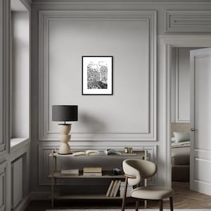 May include: A black and white illustration of a city street scene framed in a black frame. The illustration is hanging on a white wall in a room with a wooden floor and a white chair.
