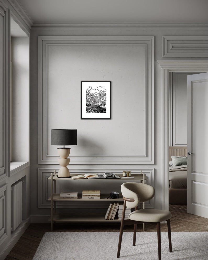 May include: A black and white illustration of a city street scene framed in a black frame. The illustration is hanging on a white wall in a room with a wooden floor and a white chair.