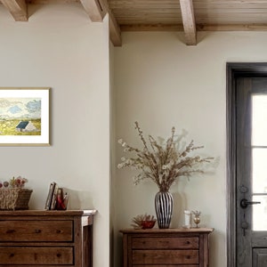 May include: A framed print of a watercolor painting depicting a small white house with a blue roof in a field. The house is surrounded by green grass and yellow flowers. The sky is blue with white clouds.