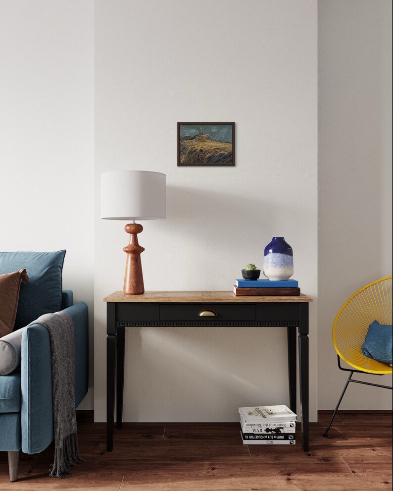 May include: A black console table with a wooden top and a single drawer. The table has a brass knob and is decorated with a framed landscape painting, a lamp, a vase with a succulent, and a stack of books.