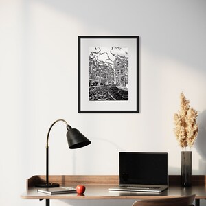 May include: Black and white line art print of a city street with buildings on either side. The print is framed in a black frame and hanging on a white wall.